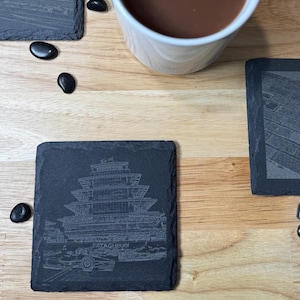 May include: Four black slate coasters with an etched design of the Indianapolis Motor Speedway. The coasters are arranged on a wooden surface with a white mug of coffee in the center.