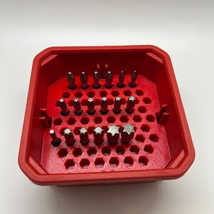 May include: A red plastic storage case with a honeycomb design holds a variety of screwdriver bits. The bits are arranged in rows and are made of metal.