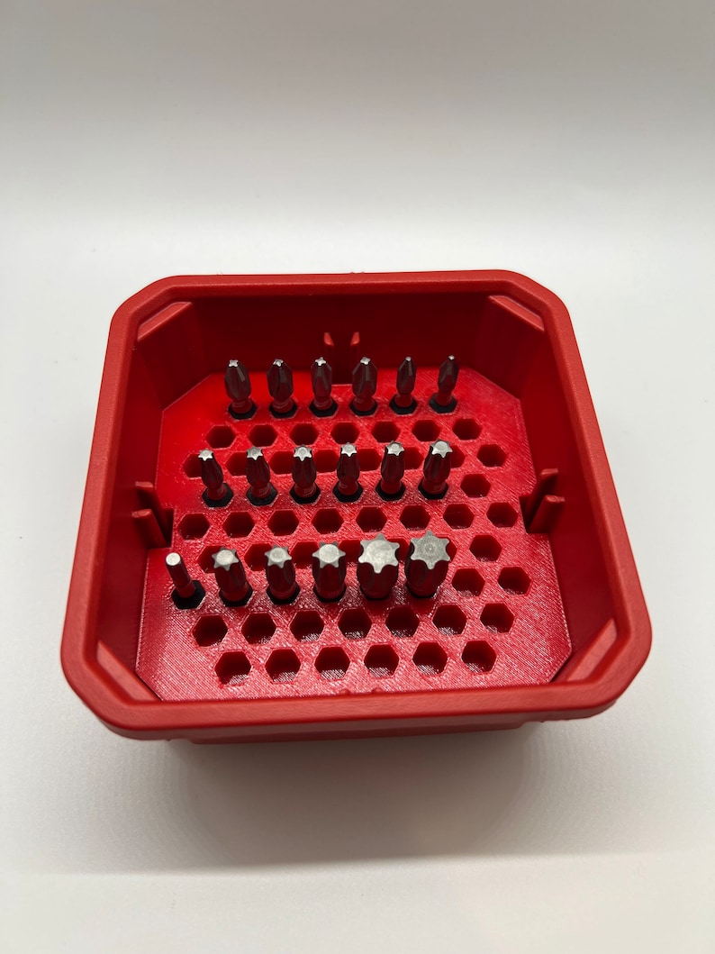 May include: A red plastic storage case with a honeycomb design holds a variety of screwdriver bits. The bits are arranged in rows and are made of metal.