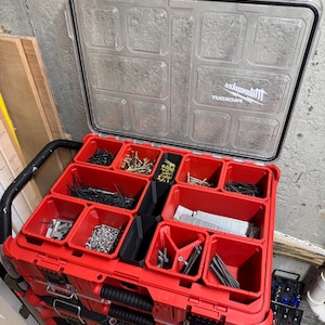 May include: A red and black Milwaukee Packout tool organizer with a clear lid, filled with various hardware items like screws, nails, and zip ties. The organizer is stacked on top of other similar units, suggesting a modular storage system.