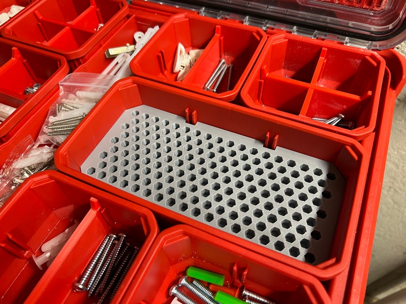 May include: A red plastic storage box with multiple compartments. The box contains a variety of small hardware items, including screws, nails, and plastic anchors. The bottom compartment has a gray honeycomb-shaped insert.