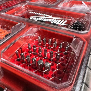 May include: A red plastic tool box with a clear plastic lid. The lid is open and reveals a variety of small metal tools, including screwdriver bits, organized in a honeycomb-shaped grid. The box has the Milwaukee Packout logo on the lid.