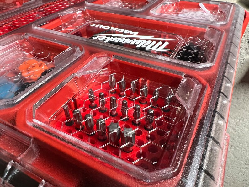 May include: A red plastic tool box with a clear plastic lid. The lid is open and reveals a variety of small metal tools, including screwdriver bits, organized in a honeycomb-shaped grid. The box has the Milwaukee Packout logo on the lid.