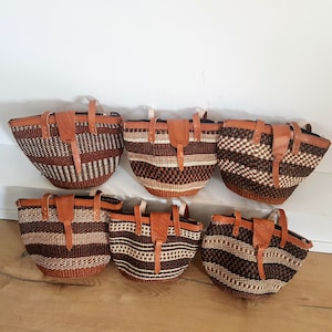 Könnte beinhalten: Sechs gewebte Körbe mit Lederdetails. Die Körbe weisen braune und beige gestreifte und gemusterte Designs auf. Jeder Korb hat einen Lederriemenverschluss und einen Griff. Die Körbe sind auf einer hellbraunen Holzoberfläche angeordnet.