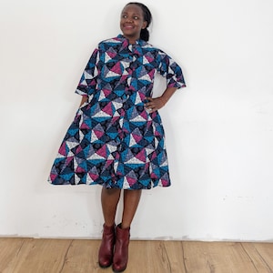 May include: A woman wearing a blue, pink and white patterned shirt dress with a button-up front. She is wearing brown ankle boots.