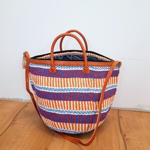 May include: A large, woven tote bag with a striped pattern in orange, blue, and white. The bag has brown leather handles and a brown leather strap.