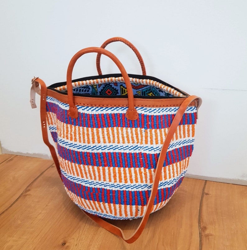May include: A large, woven tote bag with a striped pattern in orange, blue, red, and white. The bag has brown leather handles and a long shoulder strap.