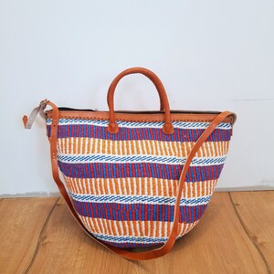 May include: A large, woven tote bag with a striped pattern in orange, blue, and red. The bag has brown leather handles and a long, adjustable shoulder strap.