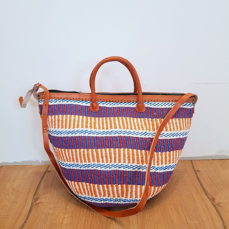 May include: A large, woven tote bag with a striped pattern in orange, blue, and red. The bag has brown leather handles and a long, adjustable shoulder strap.