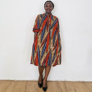Könnte beinhalten: Eine Frau trägt ein knielanges Kleid mit einem bunten geometrischen Muster in Orange, Blau und Braun. Das Kleid hat einen hohen Kragen und einen Knopfverschluss vorne.