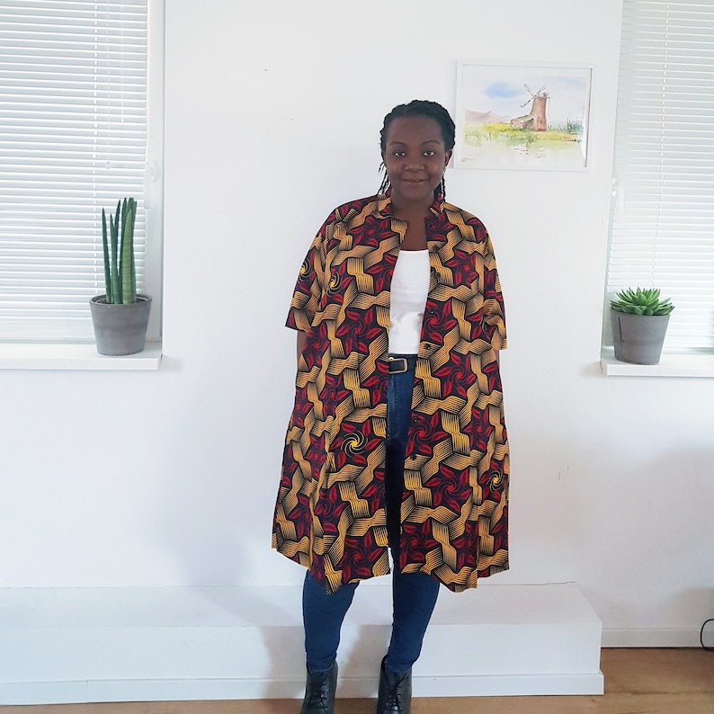 May include: A long, red, yellow, and black patterned shirt with a white collared shirt underneath. The shirt has a geometric pattern and is worn over blue jeans.