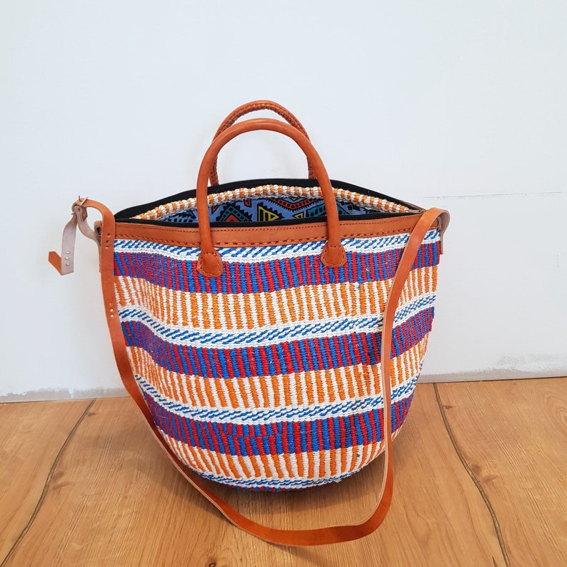 May include: A large, striped tote bag with a brown leather handle and strap. The bag is made of woven fabric and has a white, orange, blue, and red striped pattern.