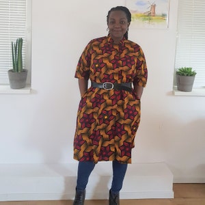 May include: A woman wearing a knee-length dress with a geometric pattern in shades of red, yellow, and black. The dress has a collared neckline and short sleeves. She is wearing a black belt with a silver buckle and black boots.