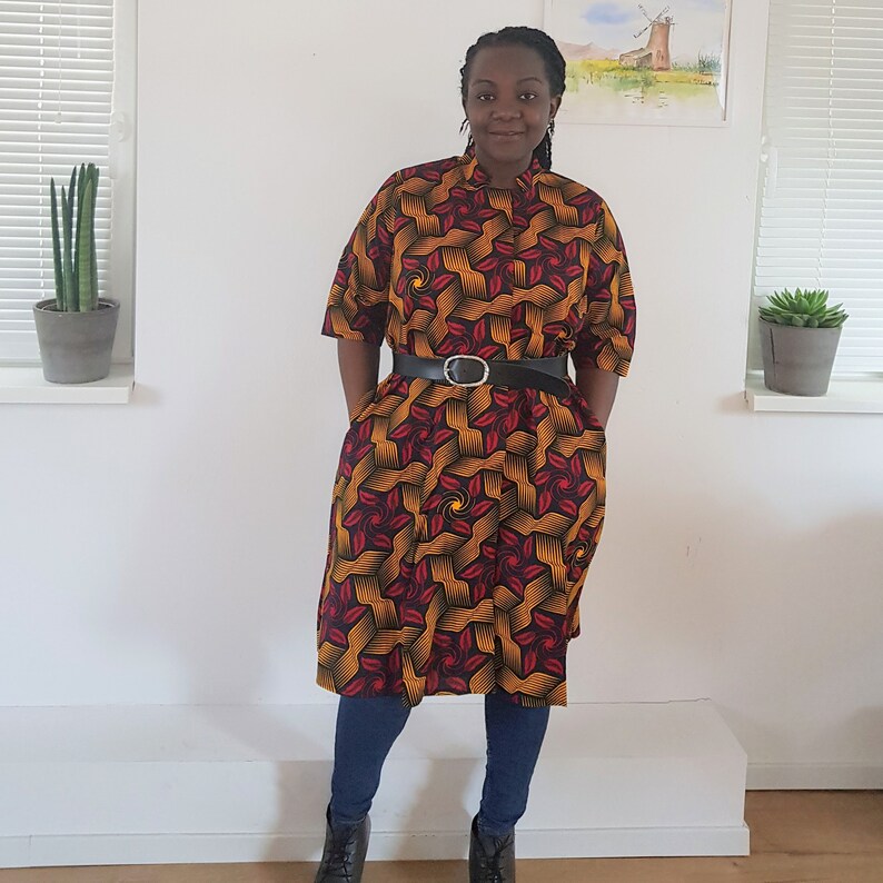 May include: A woman wearing a knee-length dress with a geometric pattern in shades of red, yellow, and black. The dress has a collared neckline and short sleeves. She is wearing a black belt with a silver buckle and black boots.
