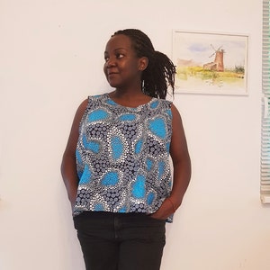 May include: A woman wearing a sleeveless top with a blue and white polka dot pattern. The top has a scoop neck and a relaxed fit. The woman is wearing black pants and has her hair pulled back in a ponytail.