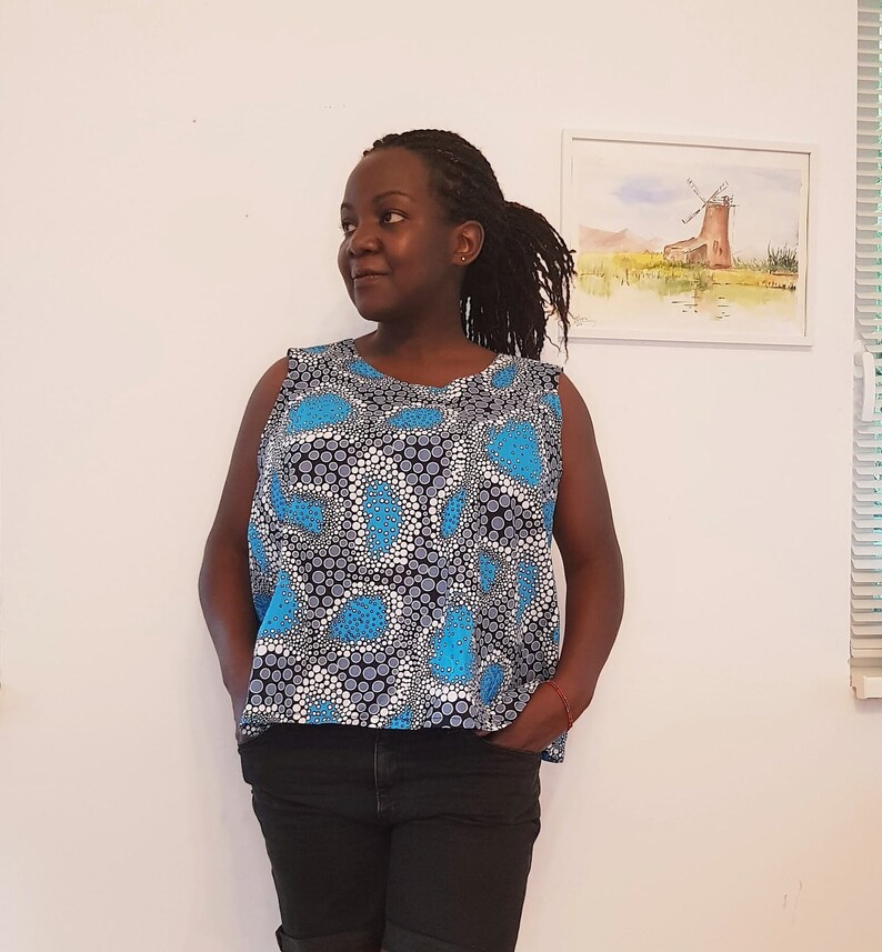 May include: A woman wearing a sleeveless top with a blue and white polka dot pattern. The top has a scoop neck and a relaxed fit. The woman is wearing black pants and has her hair pulled back in a ponytail.