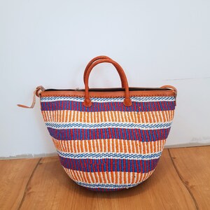 May include: A large, woven tote bag with a brown leather handle and trim. The bag is made of colorful stripes in shades of orange, blue, red, and white.