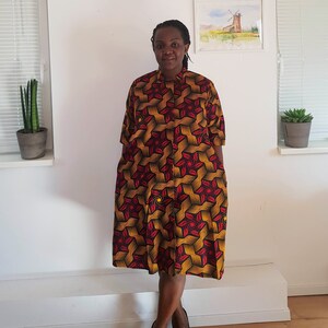 May include: A woman wearing a knee-length dress with a geometric pattern in shades of red, yellow, and black. The dress has a button-up front and short sleeves.