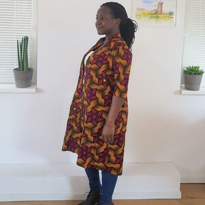May include: A long, red, yellow, and black patterned cardigan with a collar and three-quarter sleeves. The cardigan is worn over a white shirt and blue jeans.