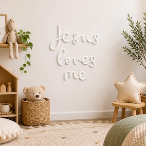 May include: White wall-mounted script lettering reading Jesus loves me, displayed above a wooden house-shaped shelf with toys, a woven basket containing a teddy bear, a small wooden stool with a star-shaped pillow, and a potted olive tree.