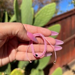 May include: Pink woven bracelet with a black eye charm and gold-colored beads. The bracelet is held in a hand with pink nail polish, with a background of green plants and a wooden fence.