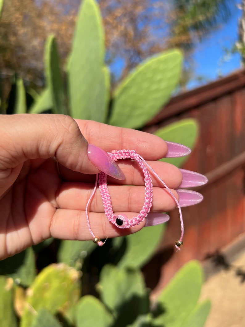 May include: Pink woven bracelet with a black eye charm and gold-colored beads. The bracelet is held in a hand with pink nail polish, with a background of green plants and a wooden fence.