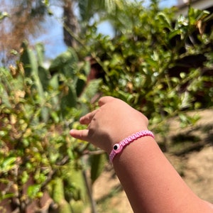 May include: A pink woven bracelet with a black and white eye charm. The bracelet is worn on a wrist, with a blurred background of green foliage and a blue sky.