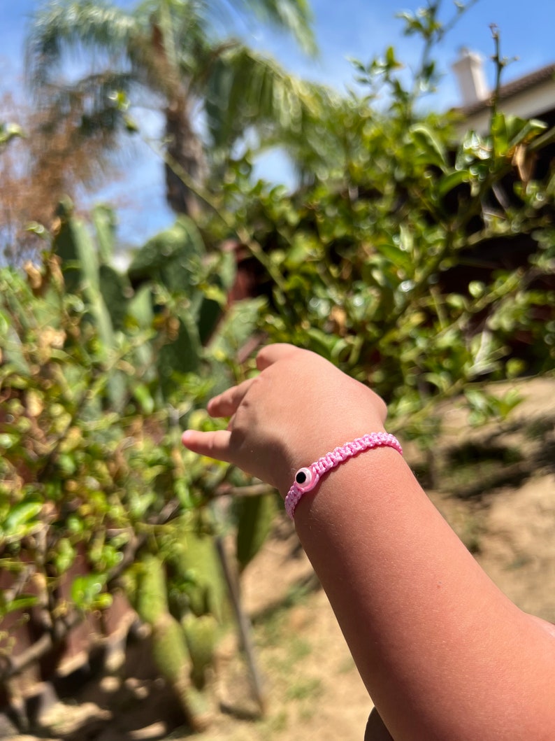 May include: A pink woven bracelet with a black and white eye charm. The bracelet is worn on a wrist, with a blurred background of green foliage and a blue sky.