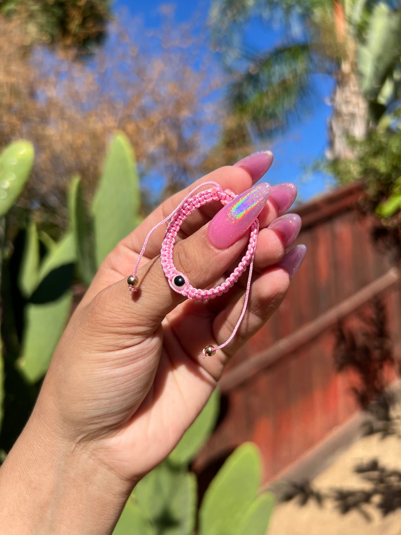May include: Pink macrame bracelet with an evil eye bead and gold-colored accents. The bracelet is held in a hand, with a blurred background of greenery and a red fence.