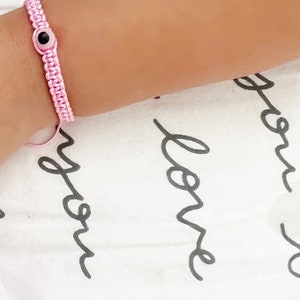 May include: A pink macrame bracelet with a black and white evil eye charm. The bracelet is on a wrist, set against a white background with black cursive text.
