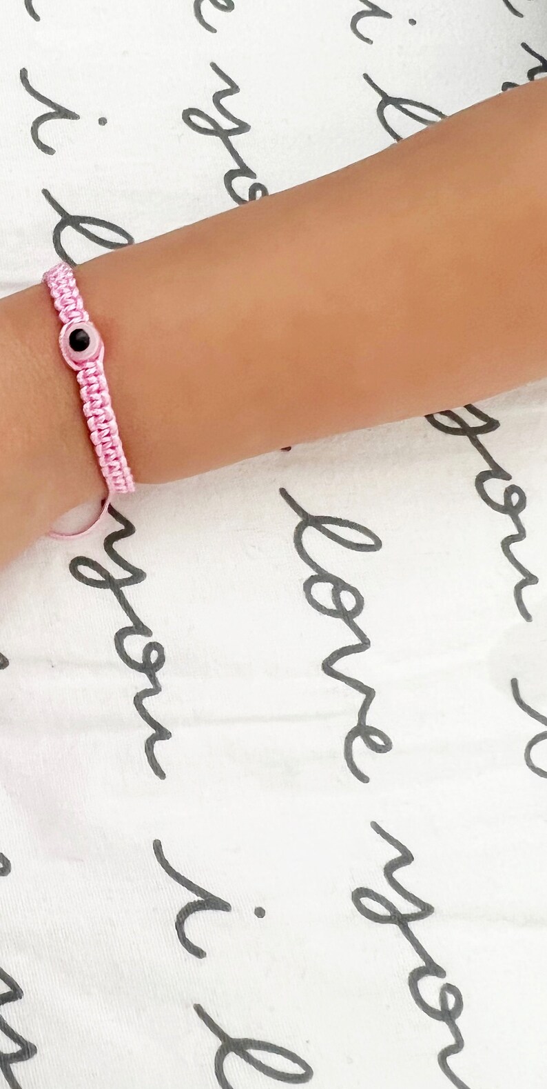 May include: A pink macrame bracelet with a black and white evil eye charm. The bracelet is on a wrist, set against a white background with black cursive text.