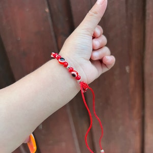 May include: Red string bracelet with small red and black evil eye beads and silver beads. The bracelet has a red string tie closure.