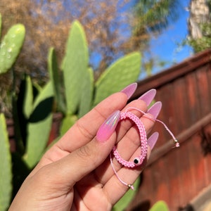 May include: Pink woven bracelet with an evil eye charm. The bracelet is made of pink thread and features a small black and white eye charm. The bracelet is held up against a background of green cactus and a blue sky.