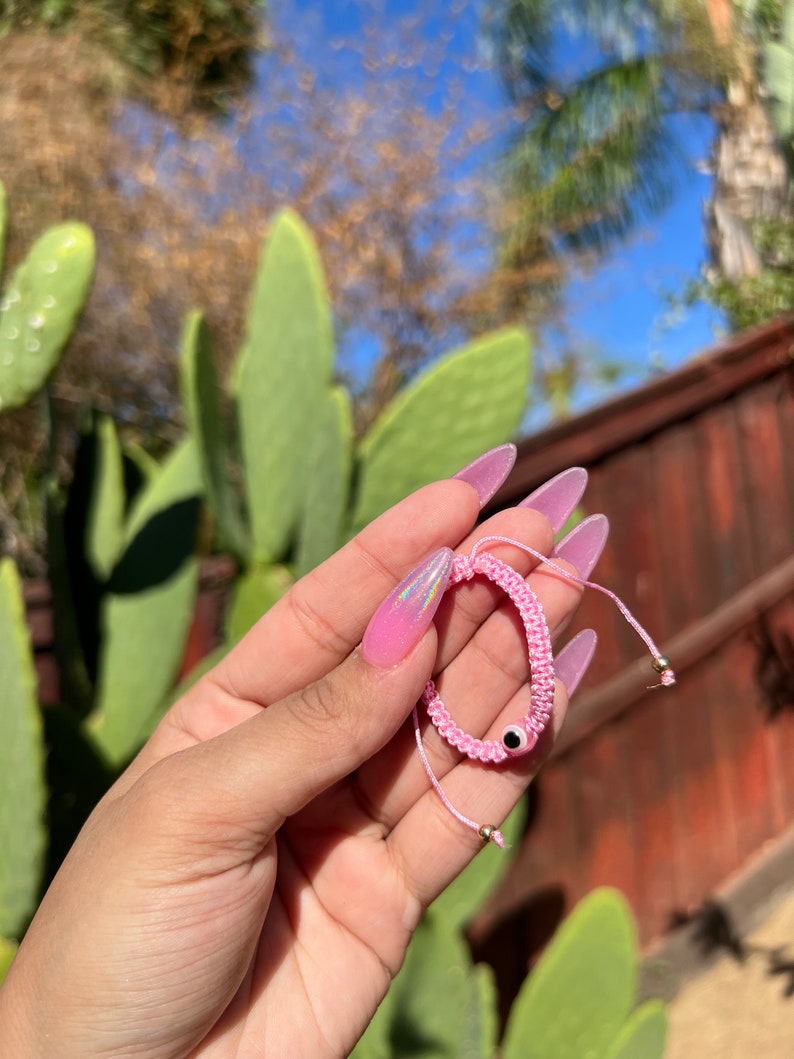 May include: Pink woven bracelet with an evil eye charm. The bracelet is made of pink thread and features a small black and white eye charm. The bracelet is held up against a background of green cactus and a blue sky.