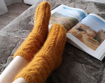 May include: A pair of mustard-yellow knitted socks with a cable knit design. The socks are resting on a gray, fluffy blanket next to an open book with landscape photographs. The socks appear soft and warm.