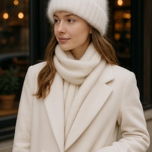 May include: A woman wearing a cream-colored winter outfit. The ensemble includes a fuzzy knit beanie, a scarf, and a long coat. The coat has a double-breasted design and a pocket. The overall look is soft and warm, perfect for cold weather.