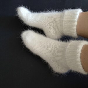 以下が含まれることがあります： リブ編みのカフが付いた白いふわふわの靴下2足。靴下は柔らかくふわふわした素材でできており、冬の足元を暖かく保つのに最適です。
