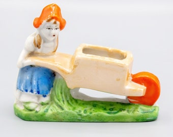 May include: A porcelain figurine of a girl in a blue dress and orange hat pushing a wheelbarrow with an orange wheel. The wheelbarrow is on a green base.