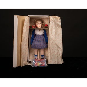 May include: A porcelain doll wearing a blue sweater, plaid skirt, and red shoes. The doll has blonde hair and is standing in a box with tissue paper.