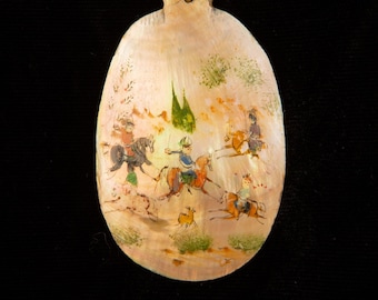 May include: A hand-painted seashell ornament depicting a scene of people riding horses in a landscape with trees and a deer. The seashell is a light pink color with a gold-colored loop at the top.