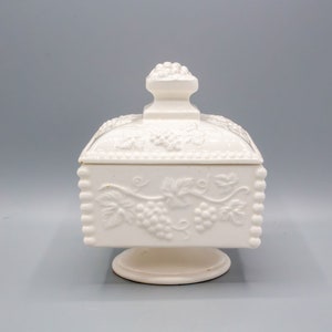 May include: White ceramic candy dish with a grapevine design and a lid. The dish has a square base and a pedestal.