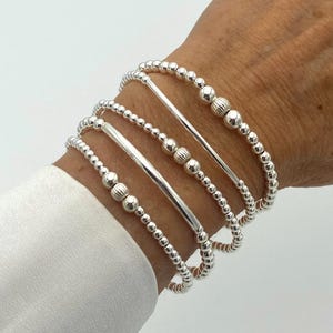 May include: A set of four silver bracelets on a wrist. The bracelets feature small, round beads and smooth, cylindrical bars. Some beads have a textured, ribbed design. The bracelets are worn against a white shirt sleeve.