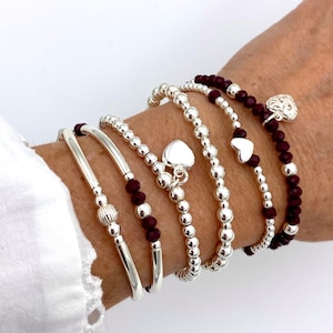 May include: A set of silver bracelets with small beads and heart charms. The bracelets are layered on a wrist and include a burgundy beaded bracelet.