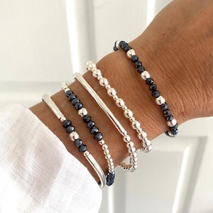 May include: A collection of silver bracelets with various designs. Some bracelets feature small, dark blue beads, while others have a smooth, metallic finish. The bracelets are stacked on a wrist, showcasing their different styles and textures.