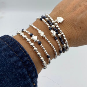 Puede incluir: Un conjunto de pulseras de cuentas plateadas con pequeños dijes de corazón plateados. Las pulseras están superpuestas en una muñeca y presentan cuentas negras.