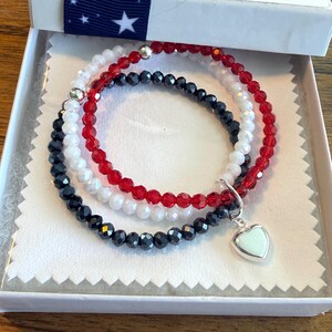 May include: A set of three beaded bracelets featuring faceted red, white, and black beads. The bracelets are accented with small silver-toned metal beads and a single silver-toned heart-shaped charm hanging from the black beaded strand.