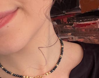 Może przedstawiać: Czarny choker z małymi złotymi koralikami i złotym wisiorem w kształcie serca. Naszyjnik nosi osoba w czarnej koszuli.