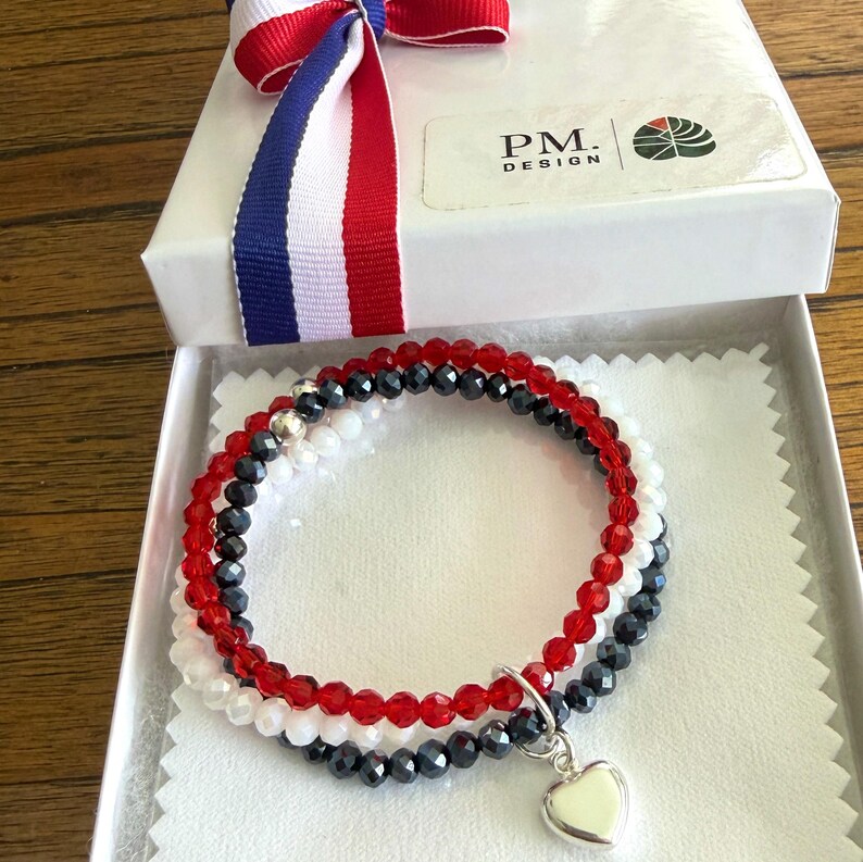 May include: A set of three beaded bracelets featuring faceted red, white, and dark gray beads. The bracelets are presented in a white gift box with a red, white, and blue ribbon and a PM. DESIGN logo. A silver heart charm hangs from the bracelets.