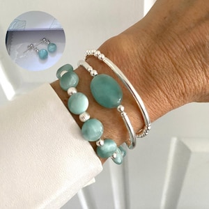 Puede incluir: Conjunto de joyas en color azul claro. La imagen muestra una pulsera con cuentas redondas y ovaladas, un brazalete de plata y pendientes de botón. Las joyas se muestran en una muñeca y una camisa blanca.