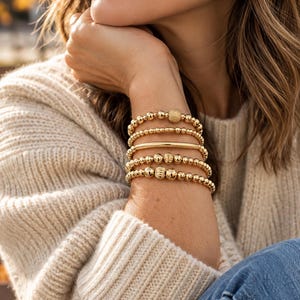 May include: A stack of five gold-toned beaded bracelets worn on the wrist. The bracelets feature a variety of round polished beads, one textured matte bead, and a single smooth horizontal bar bead, all arranged in a layered design.